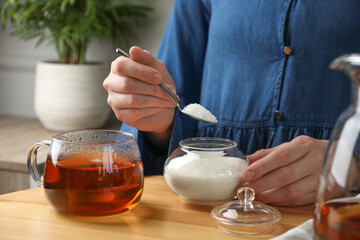 Woman adding sugar into aromatic tea at wooden table indoors, closeup