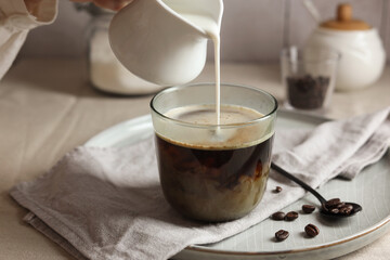 Woman pouring milk into cup with coffee at light table, closeup