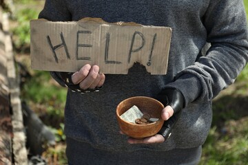 Poor homeless man holding help sign and bowl with donations outdoors, closeup