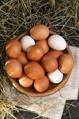 Fresh chicken eggs in bowl and dried hay on black wooden table, flat lay