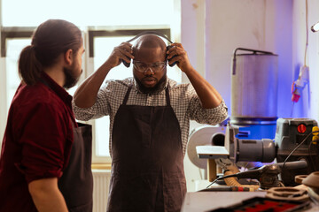 Master in carpentry shop instructing BIPOC worker to wear protection gear before assembling...