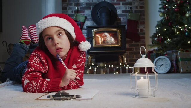 A charming little girl in a Santa hat writes a letter to Santa on the floor near the cain and Christmas tree by candlelight. Merry Christmas and Happy New Year