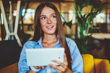 Happy young woman holding modern digital tablet and looking away while installing application for...