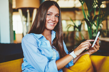 Portrait of millennial smiling hipster girl sitting at comfortable chair and using touch pad for...