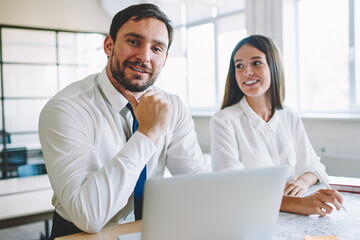 Obraz premium Portrait of successful positive male professional architect sitting at desktop with laptop computer and sketches and looking at camera while smiling female colleague near working on blueprints