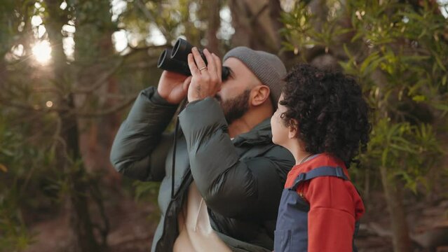 A bearded man with binoculars and a child in a forest engrossed in birdwatching together, surrounded by trees.