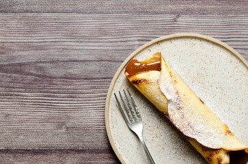 Argentinian crepe filled with dulce de leche and icing sugar on top in a beige plate on wooden background with copy space.  