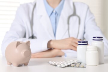 Doctor at white table indoors, focus on piggy bank and pills