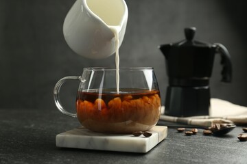 Pouring milk from pitcher into glass cup with coffee at dark textured table, closeup