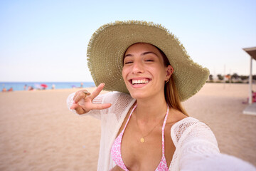 Selfie of generation z Caucasian smiling woman wearing straw hat making hand sign of peace and taking photo standing on beach. Attractive young blonde girl enjoying summer travel posing happy outdoors