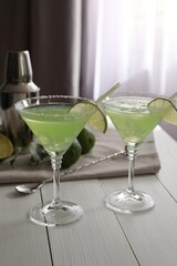 Delicious Margarita cocktail in glasses, limes, bar spoon and shaker on white wooden table