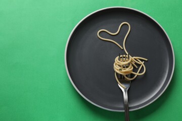 Heart made of tasty spaghetti and fork on green background, top view. Space for text
