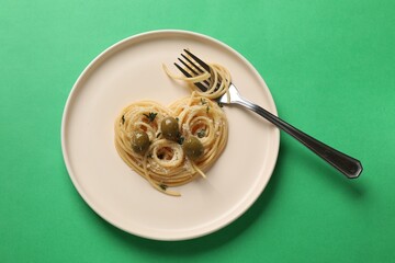 Heart made of tasty spaghetti, fork, olives and cheese on green background, top view