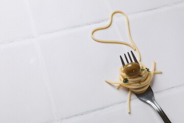 Heart made of tasty spaghetti, fork and olive on white tiled table. Space for text
