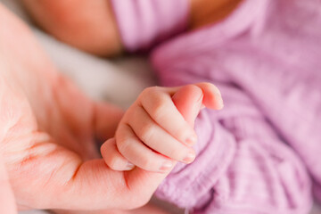 A baby wearing a purple outfit has her tiny little fingers and hand wrapped around a parent's finger.  