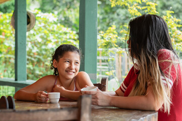 little brunette latina girl smiling while looking at her mom, mother and daughter having coffee...