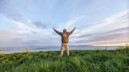 Man hiker in camo shorts and backpack on top of green hill, hands up, view on wild Atlantic way at...