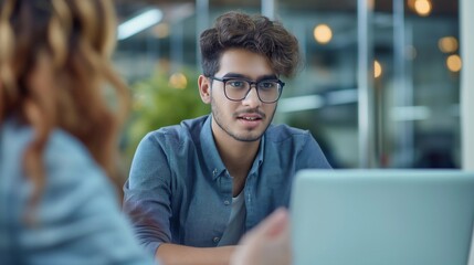 Obraz premium A young male employee with glasses, wearing business casual is sitting at a table in front of his laptop while talking to female employee about marketing data on the screen. generative AI