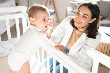 Female pediatrician measuring temperature of little baby in crib at home