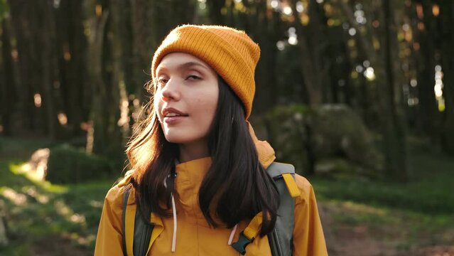 A young woman in a yellow jacket and beanie walks in a forest, looking left and right as she explores nature.
