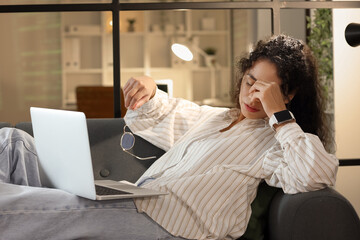 Tired female African-American programmer with laptop lying on sofa in office at night
