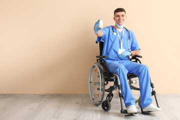 Young male doctor in wheelchair with medical mask and pills near beige wall