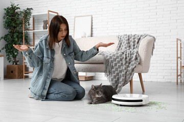 Emotional woman with cute cat, robot vacuum cleaner  and scattered litter at home