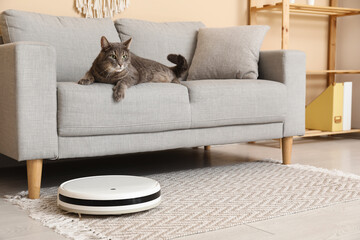 Modern robot vacuum cleaner and cute cat lying on sofa at home