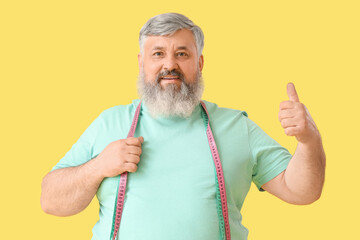 Overweight happy mature man with measuring tape showing thumb-up gesture on yellow background....