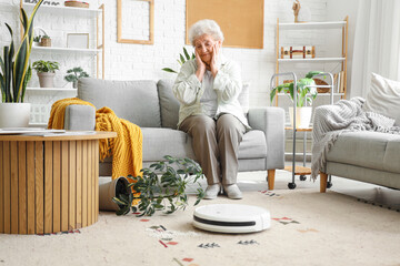 Upset senior woman with overturned houseplant and robot vacuum cleaner at home