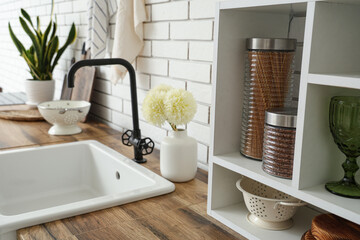 Wooden kitchen counters with sink, shelves and utensils near white brick wall