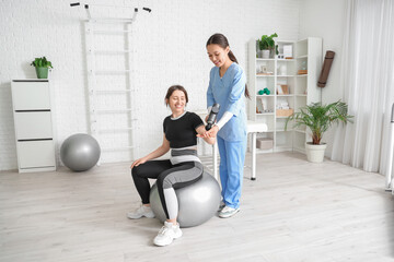Fototapeta premium Female Asian physiotherapist massaging young woman's arm with percussive massager on fitball in rehabilitation center