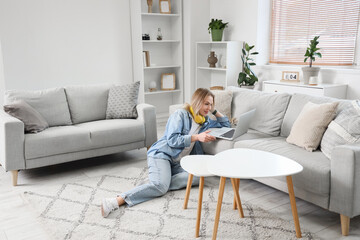 Young woman in headphones sitting on floor and using laptop on sofa in light living room
