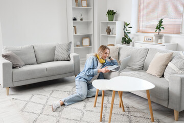 Obraz premium Young woman in headphones sitting on floor and using laptop on sofa in light living room