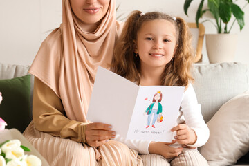Little girl with greeting card and her Muslim mother on sofa at home