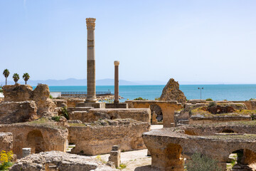 Obraz premium Sunlit Roman Baths of Antoninus ruins in Carthage on seashore, featuring remnants of marble Corinthian columns and stone structures against backdrop of blue serene waters of Mediterranean sea, Tunisia