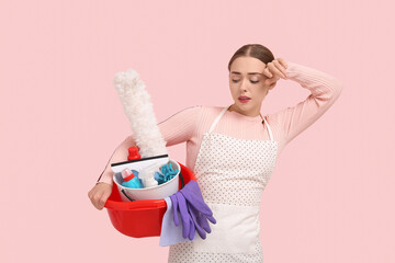 Tired young woman holding basin with cleaning supplies on pink background