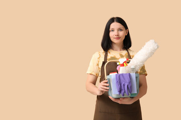 Young woman with pp-duster and cleaning supplies on beige background