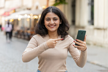 Indian woman use smartphone typing new post on web browsing searching addiction of social networks positive feedback recommends game application, showing pointing on phone. Girl on urban city street