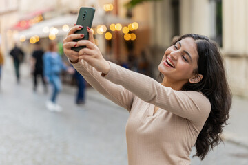Indian young woman blogger taking selfie on smartphone, communicating video call online with subscribers, recording stories for social media vlog outdoors. Hispanic girl walking on urban city street