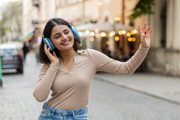 Happy overjoyed Indian young woman in wireless headphones dancing listening favorite energetic disco music in smartphone dancing outdoors. Hispanic girl walking on urban city street. Town lifestyles