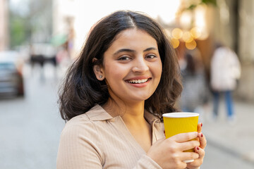 Smiling happy Indian young woman enjoying morning coffee hot drink outdoors. Relaxing, taking a break. Hispanic girl drinking coffee to go walking passes the urban city street. Town lifestyles outside