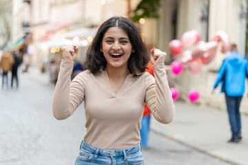 Indian young surprised woman looking at camera with big open eyes, shocked by victory, game winning, lottery goal achievement, good news outdoors. Hispanic girl walking on city street