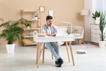 Young man using laptop at table with modern robot vacuum cleaner on floor in home office