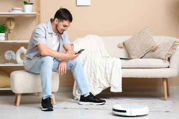 Young man with mobile phone and modern robot vacuum cleaner sitting on pouf at home