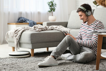 Young man in headphones listening to music with modern robot vacuum cleaner on carpet at home