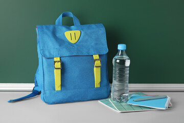 Stylish school backpack, stationery and bottle of water on table against chalkboard