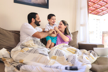 Happy family moment: parents and young son laughing together on the couch, sharing tickles and smiles while watching TV. Perfect for showcasing joyful, authentic family interactions at home.
