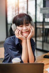 A woman wearing headphones is sitting at a desk with a laptop. She is smiling and she is enjoying her music