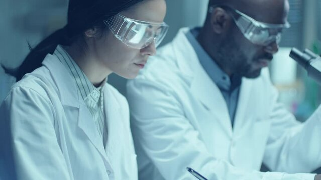 Black male chemist in protective glasses and medical coat using microscope and talking to female assistant taking notes on paper during laboratory research. Tilt-up shot